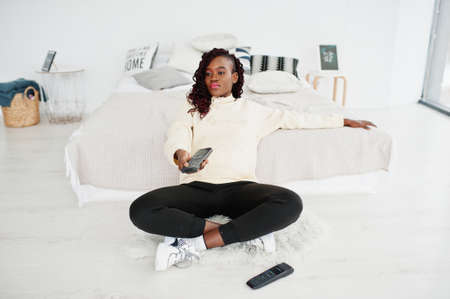 African woman watching tv at home and holding a remote control on bed.の写真素材