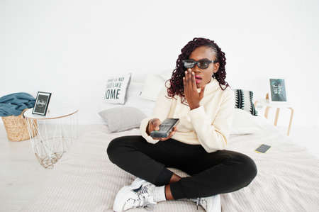 African woman in 3d glasses watching tv at home and holding a remote control on bed. Shoked face, scarry movie.の写真素材