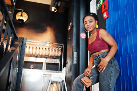 African woman in red marsala top and jeans posed indoor on stairs.