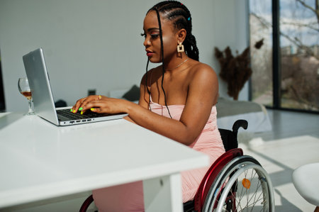 Young disabled African American woman in wheelchair at home working with laptop.