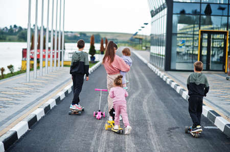 Back of young stylish mother with four kids outdoor. Sports family spend free time outdoors with scooters and skates.の写真素材