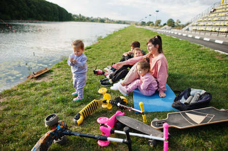 Young stylish mother with four kids sitting on grass against lake. Sports family spend free time...