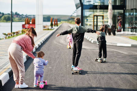 Back of young stylish mother with four kids outdoor. Sports family spend free time outdoors with scooters and skates.の写真素材