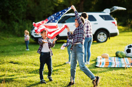 American family spending time together. Brothers play with USA flags against big suv car outdoor....