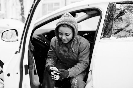 African american woman in red hoodie sitting inside car at winter snowy day with mobile phone.