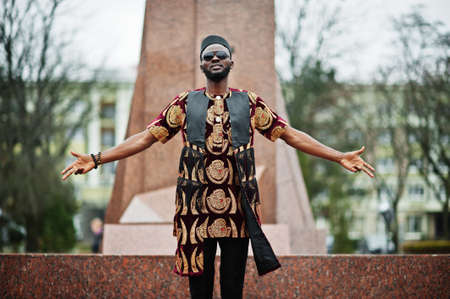 African stylish and handsome man in traditional outfit, sunglasses and black cap standing outdoor against monument.の写真素材