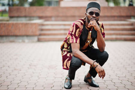African stylish and handsome man in traditional outfit and black cap squatted outdoor.の写真素材