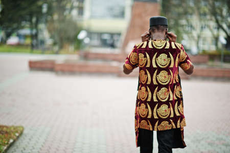 Back of african stylish and handsome man in traditional outfit and black cap standing outdoor.の写真素材