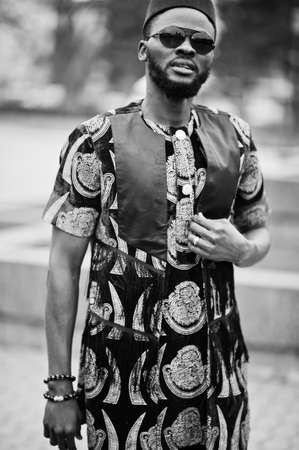 African stylish and handsome man in traditional outfit and black cap standing outdoor.