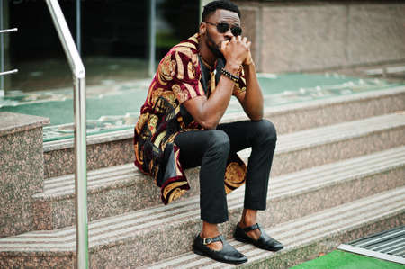 African stylish and handsome man in traditional outfit and sunglasses sitting outdoor.の写真素材
