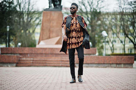 African stylish and handsome man in traditional outfit and black cap standing outdoor.の写真素材