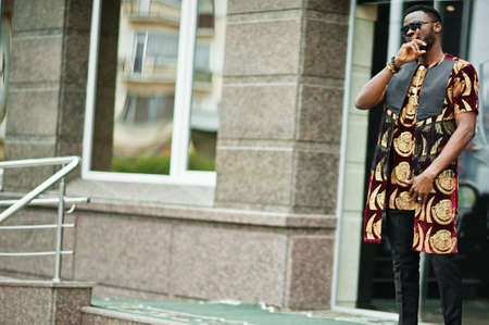 African stylish and handsome man in traditional outfit and sunglasses standing outdoor.の写真素材
