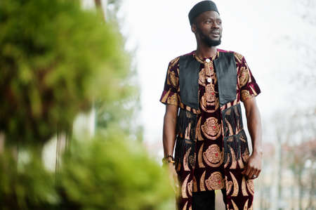 African stylish and handsome man in traditional outfit and black cap standing outdoor.