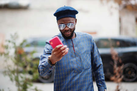 African guy in blue authentic costume, eyeglasses and beret. Fashionable nigerian man near black suv car making selfie on phone.の写真素材