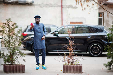 African guy in blue authentic costume, eyeglasses and beret. Fashionable nigerian man near black suv car making selfie on phone.の写真素材
