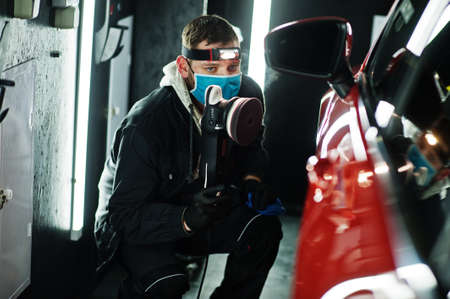 Car detailing concept. Man in face mask with orbital polisher in repair shop polishing orange suv...