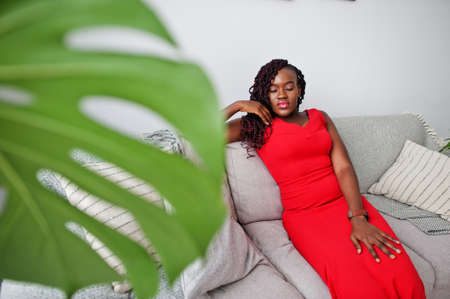 Magnificent young african woman in luxurious red dress in a luxury apartment sitting at couch. Beauty, fashion.の写真素材