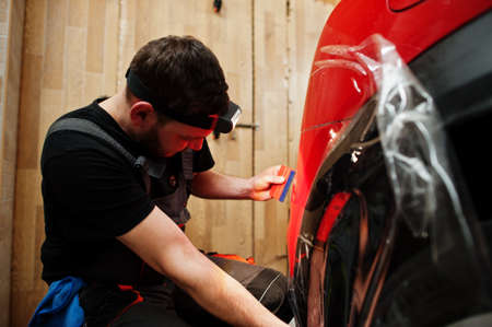 Car service worker put anti gravel film on a red car body at the detailing vehicle workshop. Car...