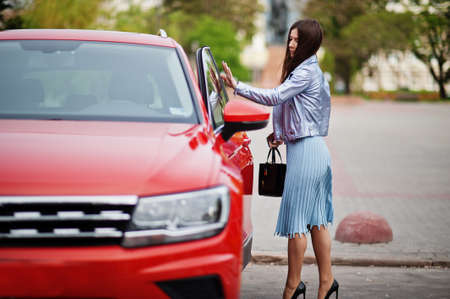 Outdoor photo of gorgeous woman with handbag posing near orange suv car.