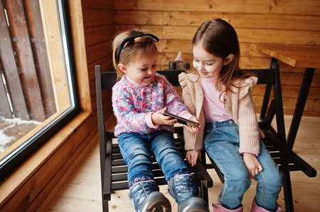 Two sisters watching at mobile phone in modern wooden house, sitting on chair.の写真素材