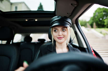 Portrait of beautiful blonde sexy fashion woman model in cap and in all black with bright makeup sit and drive red city car.の写真素材