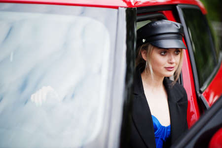 Portrait of beautiful blonde sexy fashion woman model in cap and in all black, blue corset, with bright makeup near red city car.の写真素材