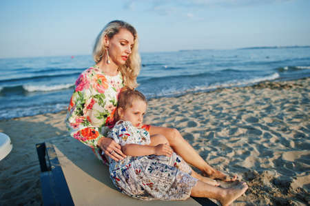 Mother and beautiful daughter having fun on the beach, sitting on sunbed. Portrait of happy woman with cute little girl on vacation.の写真素材