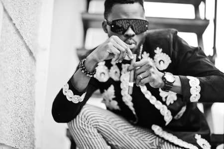 Mega stylish african man in traditional jacket pose. Fashionable black guy in sunglasses with cigar.