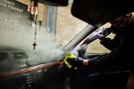 Man in uniform and respirator, worker of car wash center, cleaning car interior with hot steam...