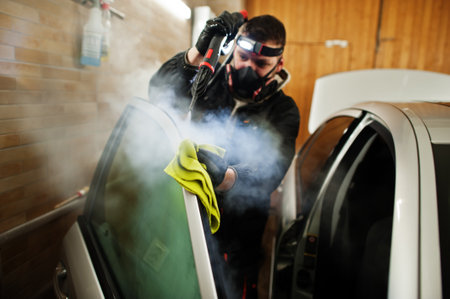 Man in uniform and respirator, worker of car wash center, cleaning car interior with hot steam cleaner. Car detailing concept.の写真素材