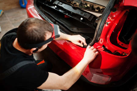 Car service worker put anti gravel film on a red car body at the detailing vehicle workshop. Car protection with special films.の写真素材