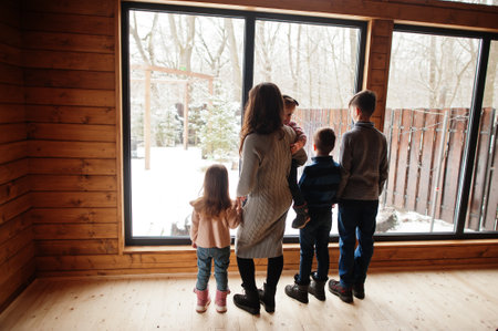 Mother and four kids in modern wooden house against large window.