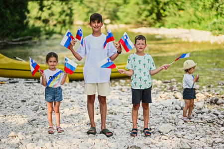Four kids hold slovenian flags in rocky shore of a calm river in Triglav National Park, Slovenia.