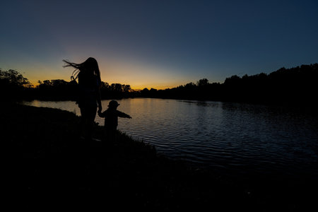 Silhouettes of mother and daughter holding hand against lake in sunrise dawn.の写真素材