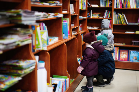 Little cute baby in jacket reaching a book from bookshelf at the library. Learning and education of...