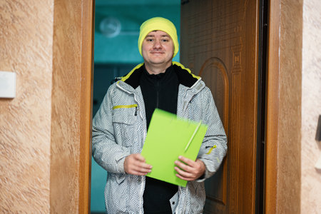 Delivery man. Courier in green color hat holding a clipboard while standing against door of...