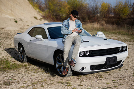 Handsome man in jeans jacket and cap sit on hood at his white muscle car in career and texting on...