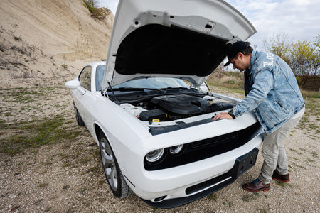 Handsome man in jeans jacket and cap is standing near his white muscle car with opening hood, check...