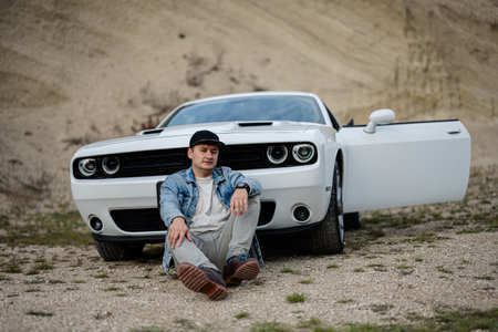 Handsome man in jeans jacket and cap sitting near his white muscle car in career.