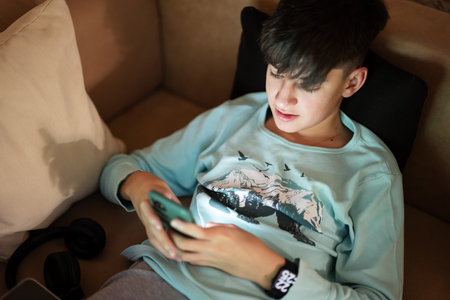 Young teen boy texting phone in front of a laptop on a bed at evening.の写真素材