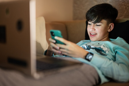 Young teen boy texting phone in front of a laptop on a bed at evening.