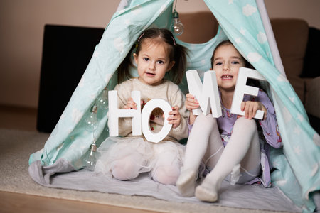 Two girls sisters at wigwam tent with wooden home letters.の写真素材
