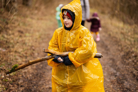 Boy in yellow raincoat with log at hands, against mother and children in the forest after rain in raincoats together.の写真素材