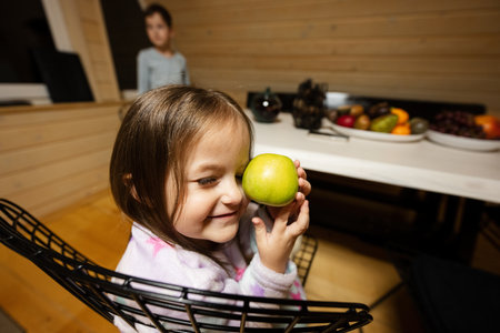 Little cute baby girl in pajama sit kitchen with green apple in cozy wooden tiny cabin house. Life in countryside.の写真素材