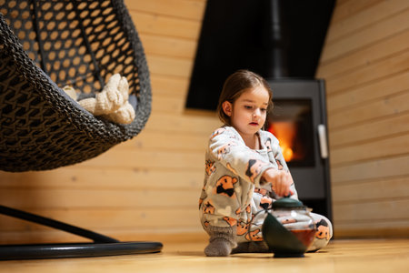 Baby girl in pajama and knitted socks with teapot against fireplace in cozy wooden tiny cabin house. Life in countryside.の写真素材