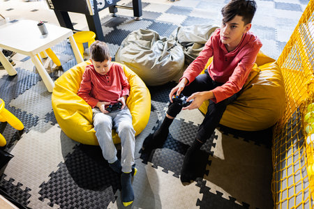 Two brothers playing video game console, sitting on yellow pouf in kids play center.