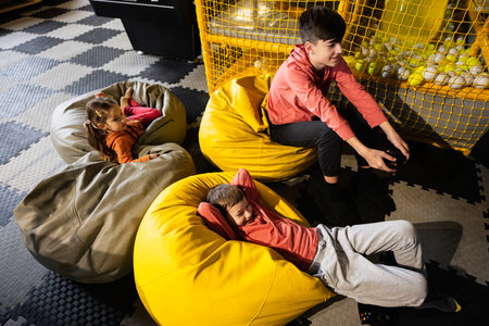 Three children playing video game console, sitting on yellow pouf in kids play center.の写真素材