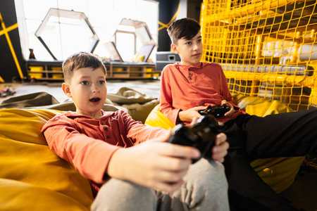 Two brothers playing video game console, sitting on yellow pouf in kids play center.の写真素材