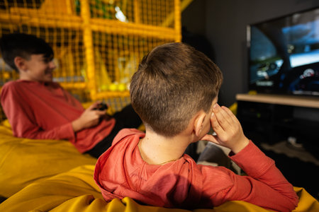 Two brothers playing race video game console, sitting on yellow pouf in kids play center.の写真素材