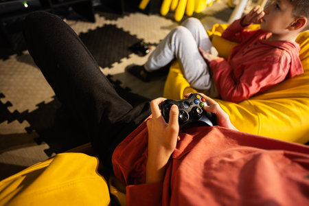 Two brothers playing video game console, sitting on yellow pouf in kids play center.の写真素材